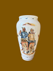 A cream-colored clay vase with hand-carved and hand-painted designs featuring two figures on horseback. The vase sits against a wooden background with a red metal object in the background.